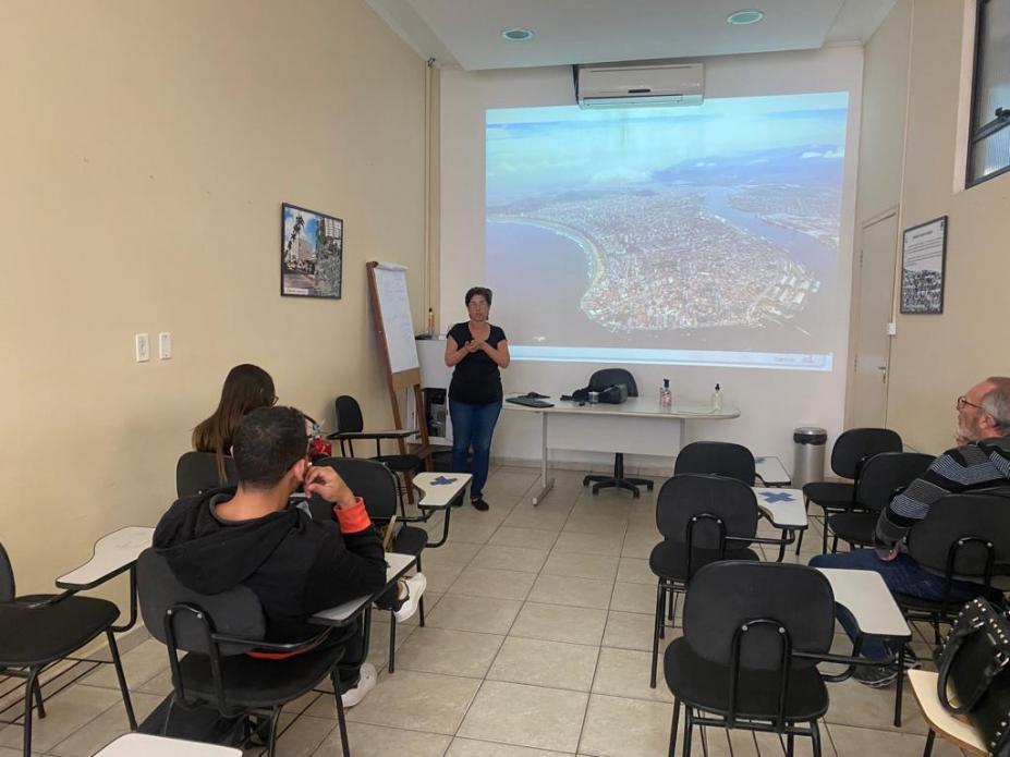 pessoas sentadas em sala de aula observam palestrante com telão ao fundo #paratodosverem