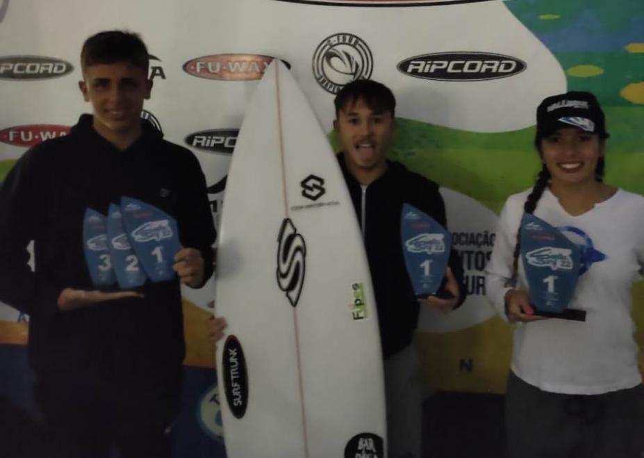 tres atletas segurando troféus e o do centro segurando prancha também #paratodosverem