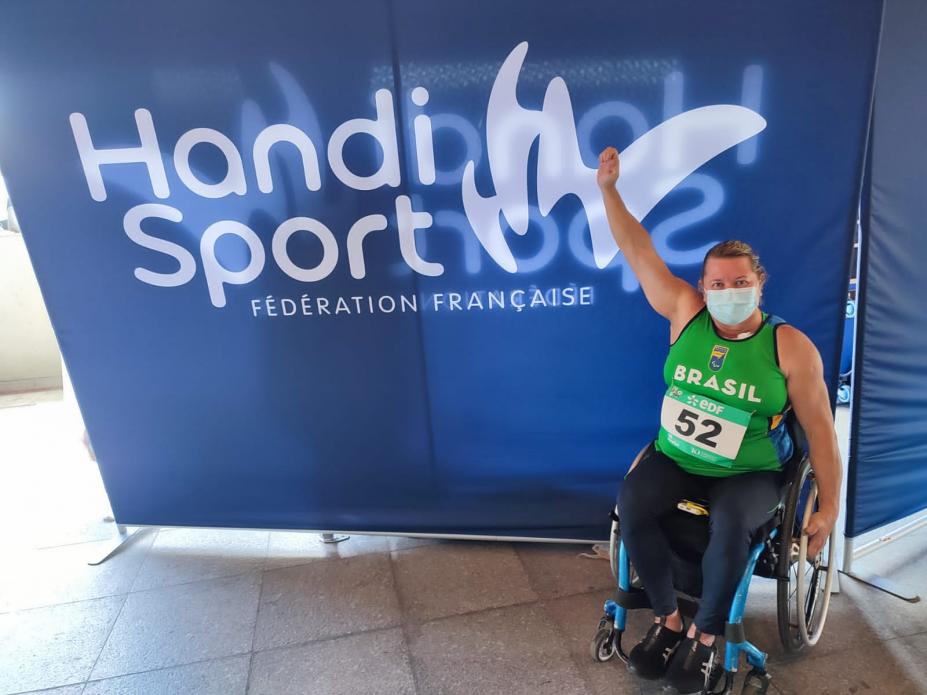 atleta na cadeira de rodas levanta o braço com banner do evento ao fundo #paratodosverem