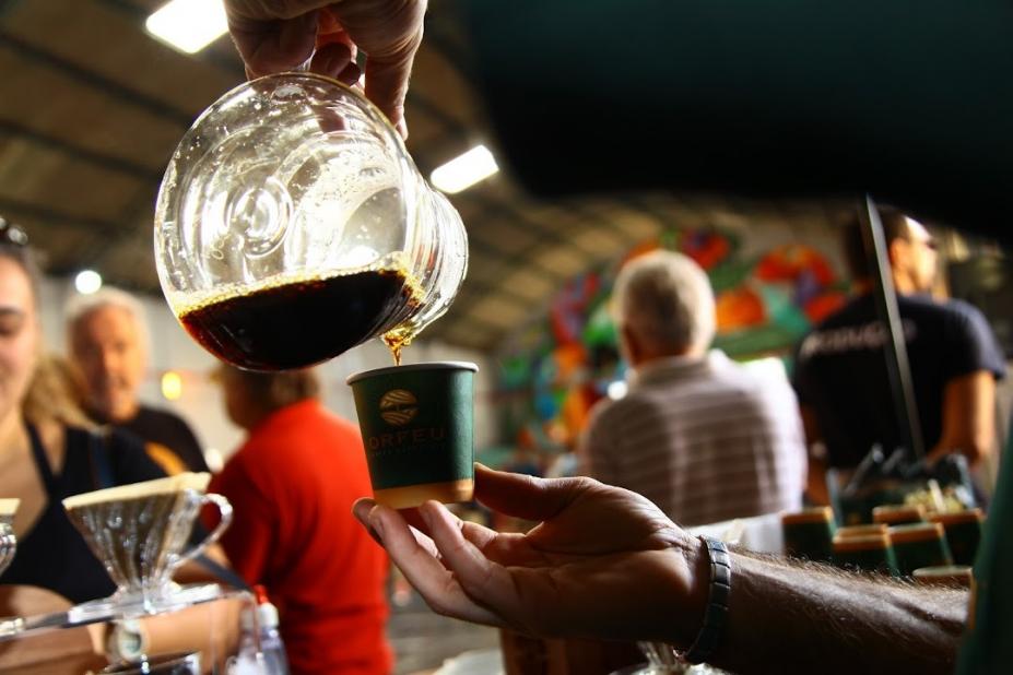 imagem mostra café em jarra sendo servido em copo com pessoas ao fundo #paratodosverem