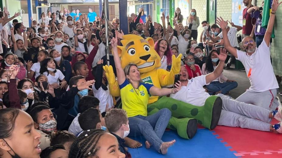 alunos professores e mascotes sentados com a mão levantada #paratodosverem