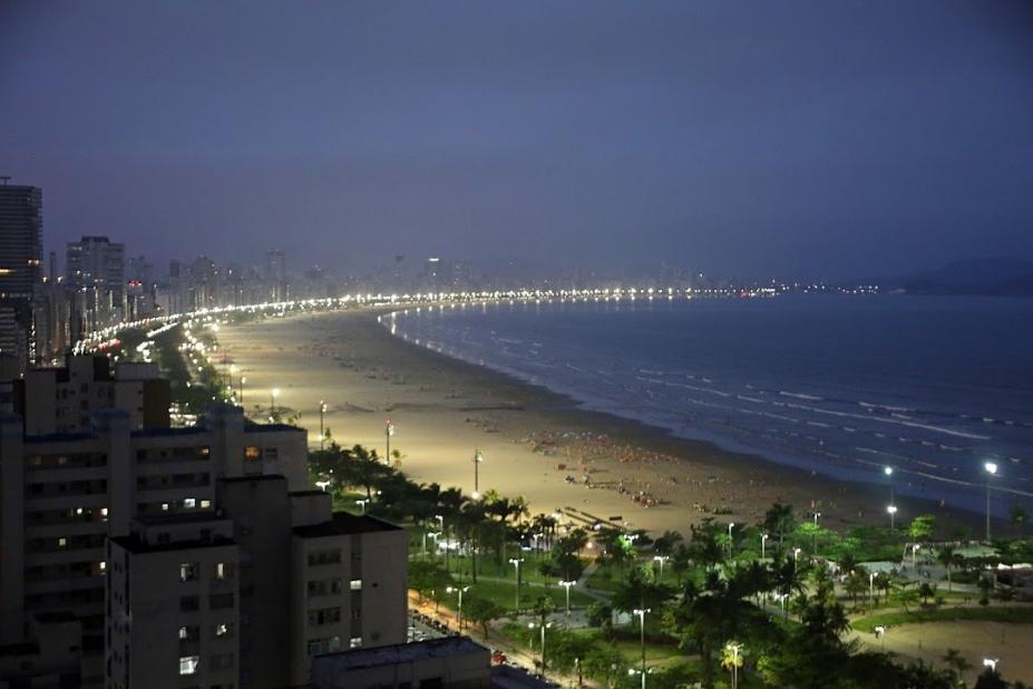 Imagem área da praia a noite, aparecendo prédios, jardins, faixa de areia e mar #paratodosverem