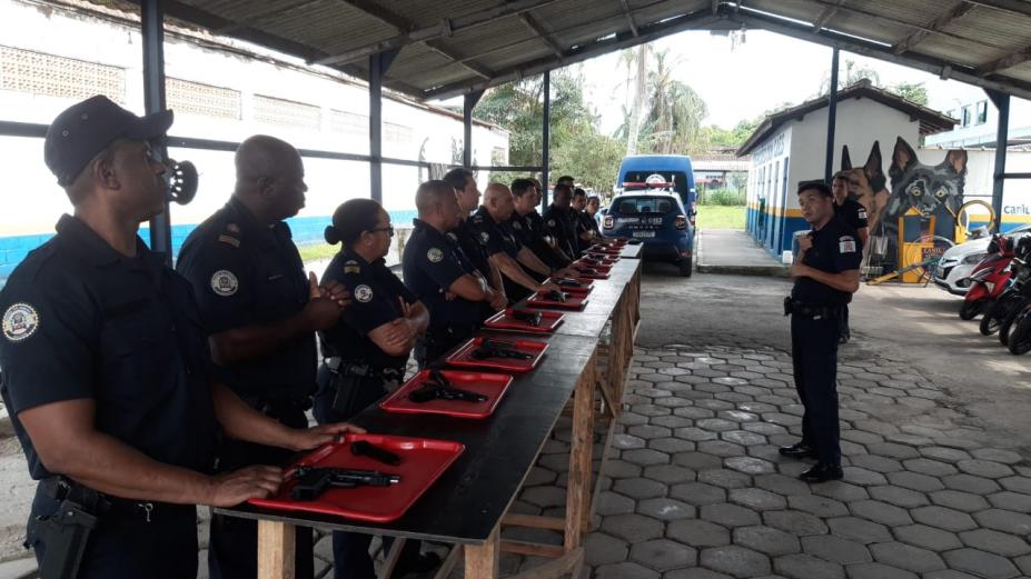 guardas municipais estão em pé atrás de bancada. À frente de todos há bandejas com arma e munição. #paratodosverem