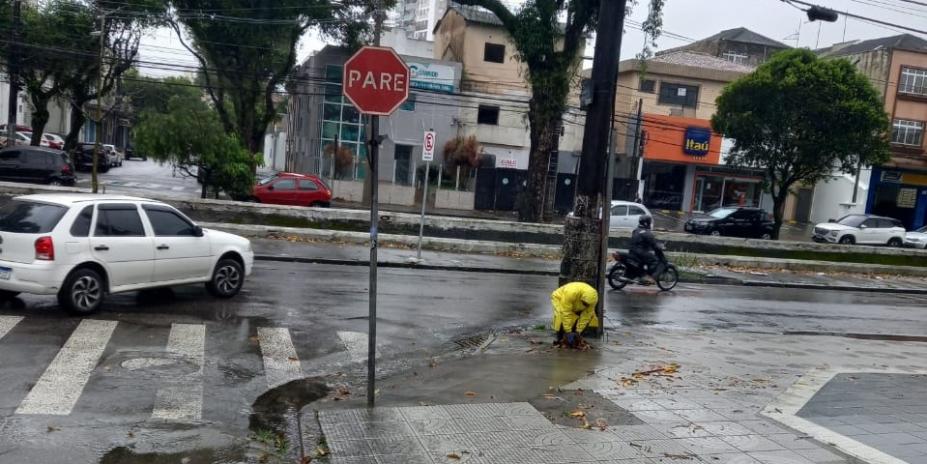 agente usando capa de chuva está junto a bueiro finalizando desobstrução. #paratodosverem