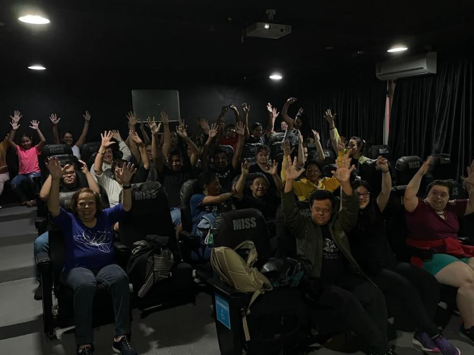 pessoas com a mão para cima sentadas em cadeiras em sala de cinema #paratodosverem