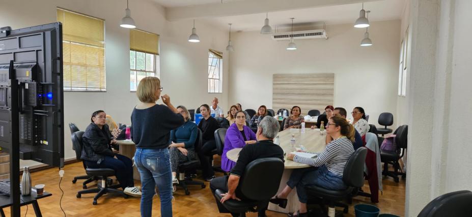 Reunião de várias pessoas que estão sentadas em torno de uma mesa e uma mulher, de costas para a foto, falando ao público. #paratodosverem