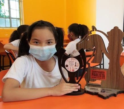 Elisa posa para foto, sentada e ao lado de troféu sobre a mesa. #paratodosverem