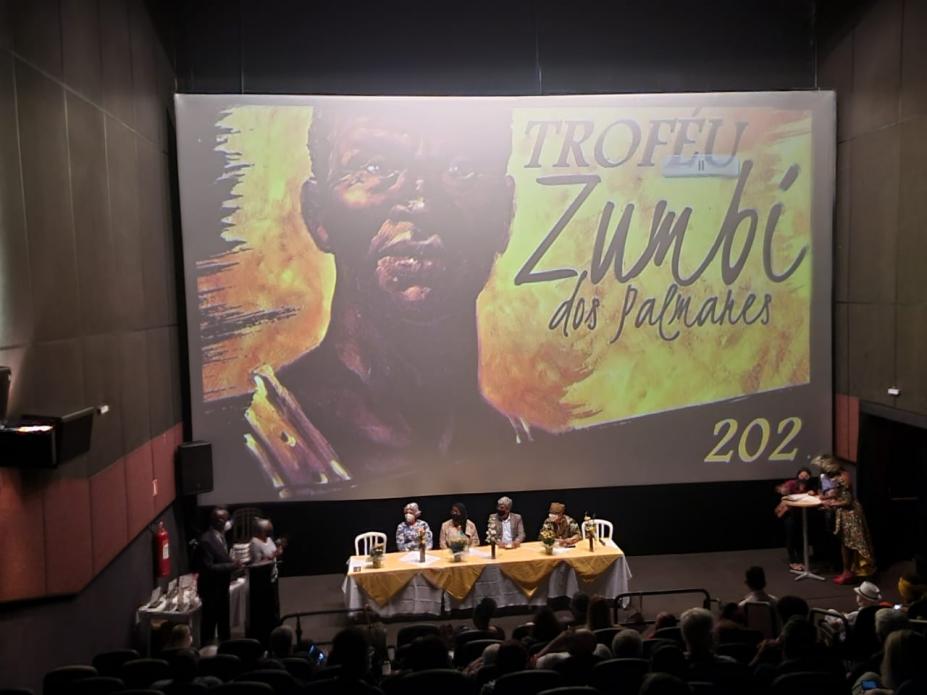 No cinema, telão com imagem de Zumbi dos Palmares e abaixo mesa com autoridades e platéia #paratodosverem