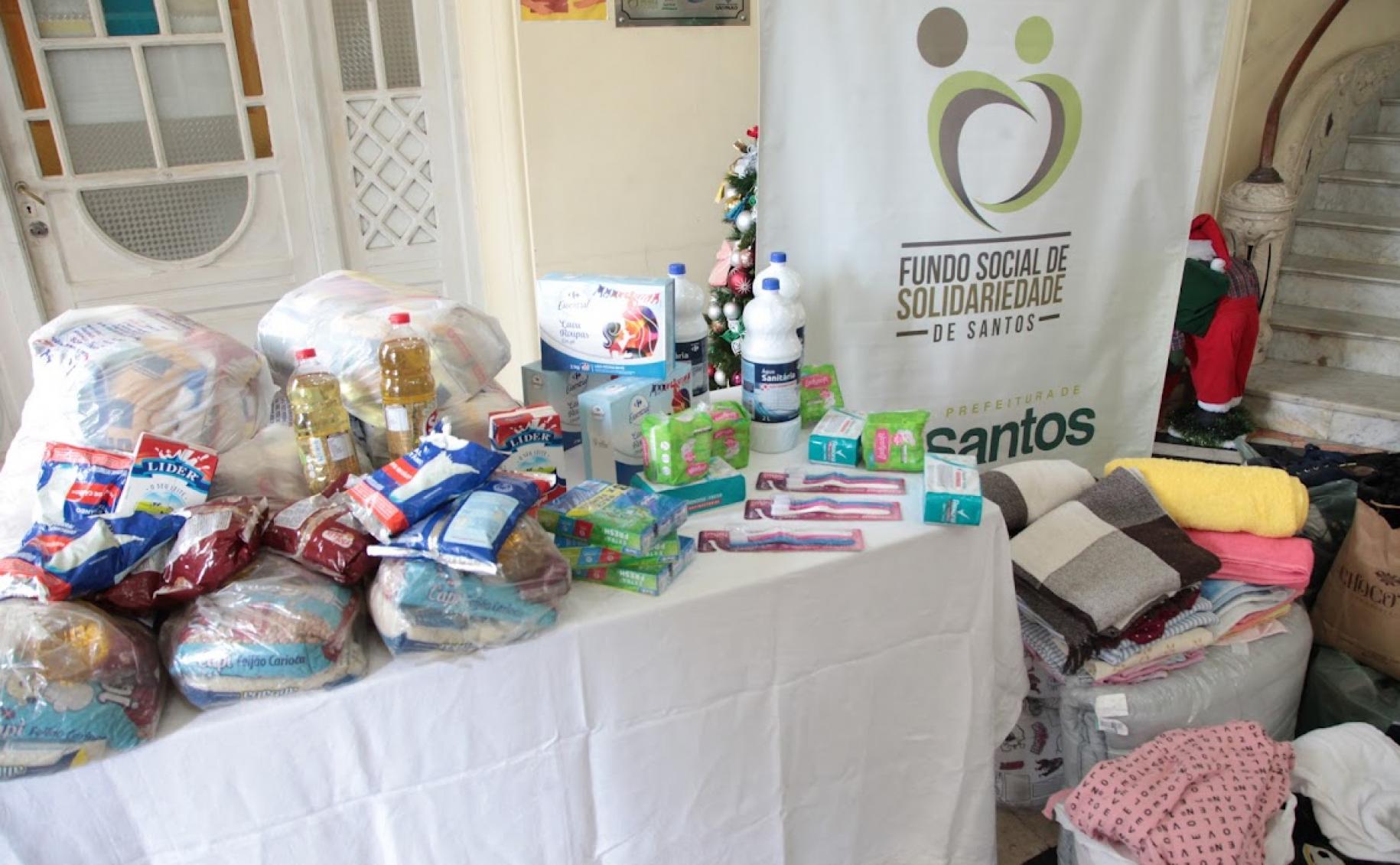 mesa com várias doações de alimentos e roupas empilhadas ao lado #paratodosverem