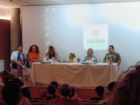 mesa de debates com seis pessoas sentadas e Wilma ao centro. #paratodosverem