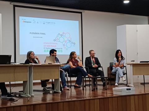 cinco pessoas sentadas diante de mesa e à frente de telão. #paratodosverem
