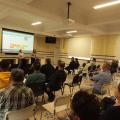 várias pessoas sentadas em um salão assistindo palestra. Ao fundo há uma imagem projetada em telão. #paratodosverem