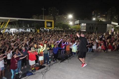 Evento é um convite para quem já sabe dançar, quer aprender ou só se divertir público dança diante de palco animado por instrutor. #paratodosverem