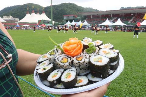 Undokai é marcado por muita gastronomia oriental, apresentações culturais, atividades recreativas e gincanas ao ar livre. Fotos: Raimundo Rosa prato de comida japonesa com dança ao fundo #paratodosverem