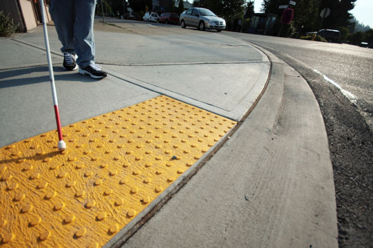 À esquerda, bengala de cego 'toca' o piso tátil amarelo de uma calçada em concreto desempenado. Ao fundo, vê-se parte do corpo de uma pessoa. À direita, uma rua, com um carro ao fundo. #pracegover