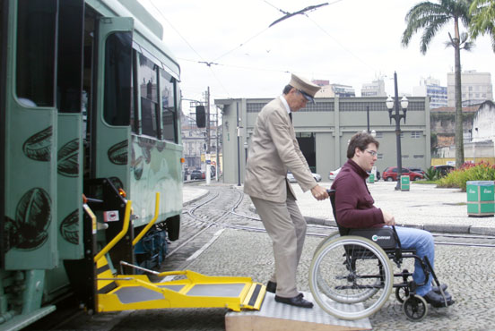 Na imagem é possível ver o elevador e rampa adaptados do Bonde Café e motorneiro auxiliando cadeirante a sair do veículo. #pracegover