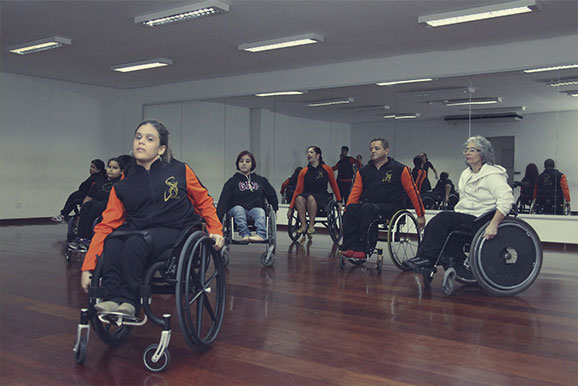 Sete cadeirantes, sendo uma idosa, um homem e cinco jovens, todos brancos, e a maioria com uniformes parecidos, na cor preta e vermelha, estão em suas respectivas cadeiras de rodas, espelhado por um salão. Ao fundo, parede envidraçada. #pracegover