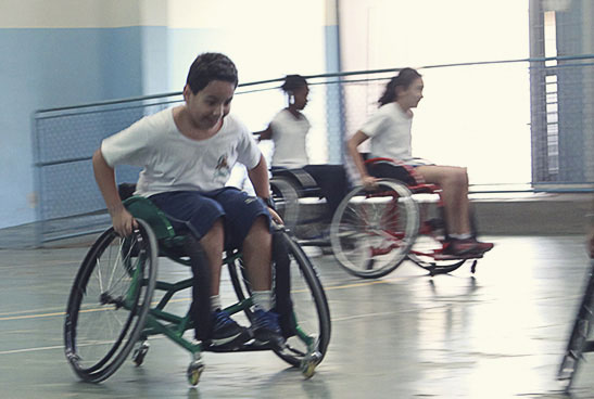 Em primeiro plano, menino cadeirante branco, uniformizado, faz movimento de ação com sua cadeira de rodas. Ele está sorrindo. Ao fundo, desfocado, duas jovens cadeirantes, uma negra e outra branca, em movimento. Atrás delas, há uma rampa acessível. #pracegover