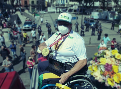 Jovem branca sorridente, vestida com maiô vermelho, onde se lê: 'Fupes', está com o braço, semi amputado, levantado. Nele, estão penduradas diversas medalhas, com cordões nas cores preta, vermelha e branca. #pracegover