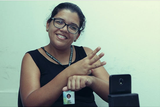 Mulher branca de óculos conversa com homem branco de óculos, de costas, em linguagem de sinais. Os dois estão sentados numa mesa com um computador. Ao fundo, em cartaz preso na parede, lê-se: 'Central de Interpretação de Libras', com letras em tonalidade verde e azul. Logo abaixo, há três logomarcas: Condefi, Prefeitura de Santos e Governo Federal. #pracegover