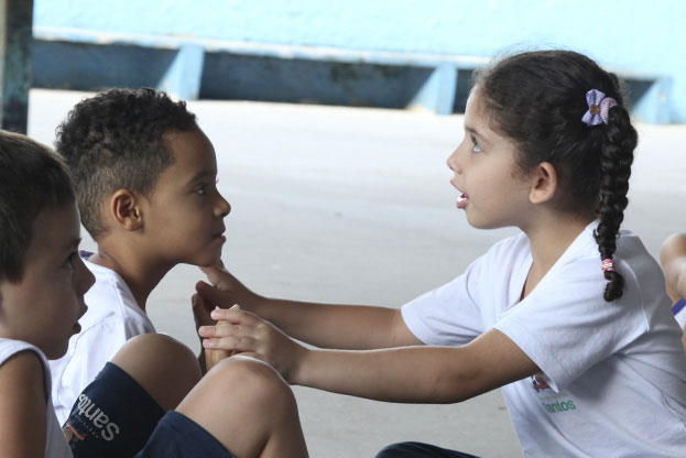 Dois meninos, um branco e um negro, sentados e uniformizados, observam a ação de uma menina branca, também sentada, em frente à eles, com a boca semi cerrada e os olhos virados para cima. Fundo desfocado. #pracegover