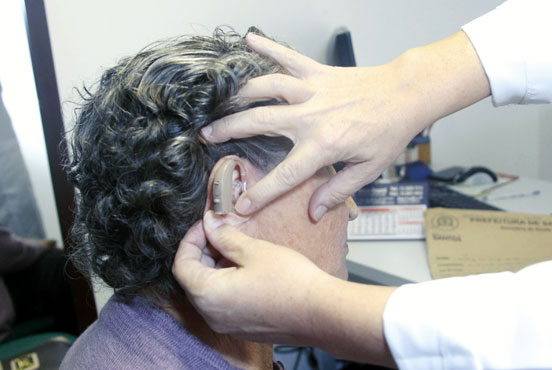 Mãos de médica, de jaleco branco, estão tateando aparelho de surdez, instalado no ouvido de uma idosa. A idosa está sentada, com o rosto virado para trás. Ao fundo, mesa com papéis, um envelope pardo e equipamentos médicos. #pracegover