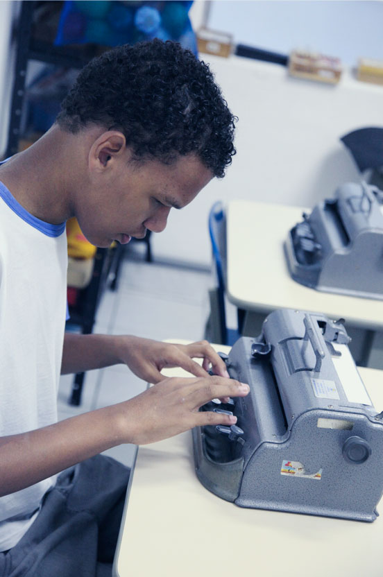 Jovem negro, sentado e com a cabeça curvada para baixo, vestido com camiseta branca, digita em máquina de escrever para cegos. O aparelho está numa bancada. #pracegover