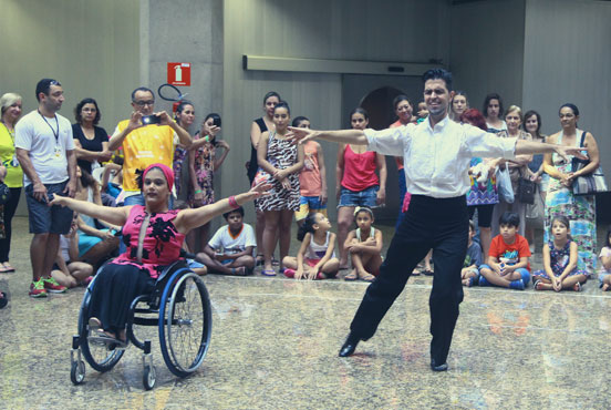 Jovem cadeirante branca, com turbante e roupa rosa, está ao lado de homem branco, em pé, com camisa branca e calça preta. Ambos estão com os braços levantados até a altura da cabeça, num movimento de dança. Ao fundo, dezenas de crianças e adultos, alguns sentados e outros em pé, observam a ação. No meio deles, um homem fotografa os dançarinos. #pracegover