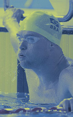 Foto em tom azulado. Rosto de homem branco, dentro de um piscina, com uma da mãos na borda. Ele usa óculos branco para natação e toca branca. Na toca está estampada a bandeira brasileira e, logo abaixo, lê-se: 'Bra'. #pracegover