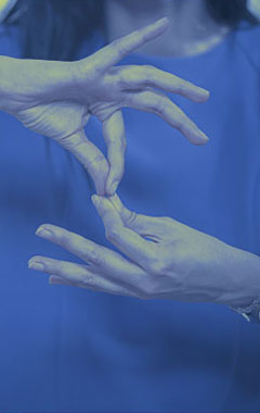 Foto em tom azulado. No centro da imagem, duas mãos simulam a linguagem de sinais para surdos. #pracegover
