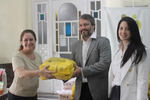A representante do Fundo Social, o head de novos negócios da empresa, Frederico Moura, e a gerente de novos negócios da empresa, Frederico Moura mulher e homem seguram pacote de cesta básica ao mesmo tempo. Ao lado do homem há outra mulher. Todos sorriem para a foto. #paratodosverem